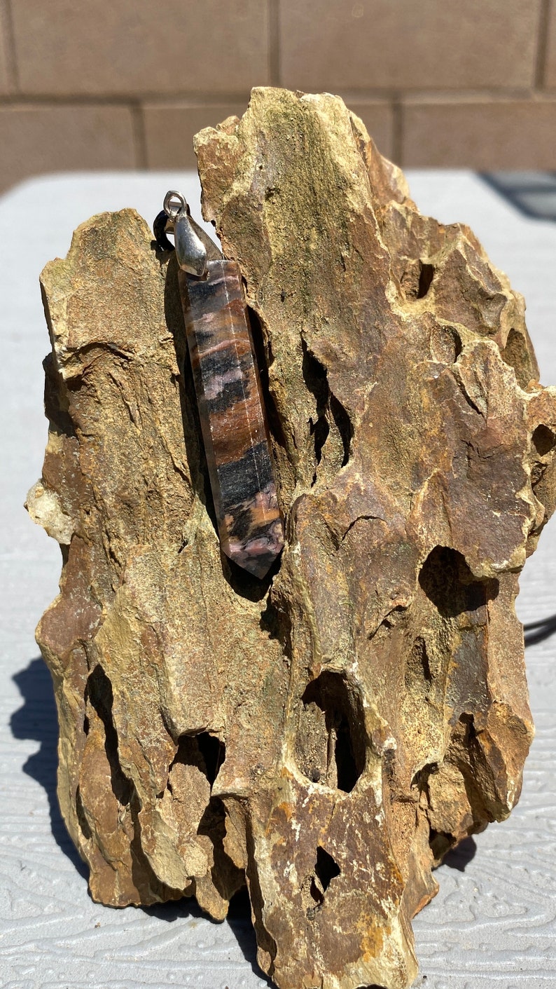May include: A silver pendant with a long, pointed, brown and black striped crystal. The crystal is set in a silver setting and is hanging from a silver chain. The pendant is resting on a rough, brown rock.
