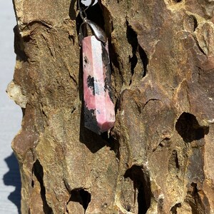 May include: A pink and black crystal pendant with a silver setting. The pendant is hanging from a black cord and is resting on a piece of rough, brown rock.