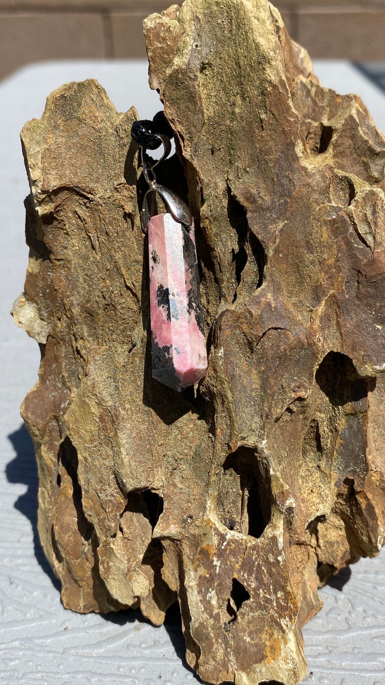 May include: A pink and black crystal pendant with a silver setting. The pendant is hanging from a black cord and is resting on a piece of rough, brown rock.