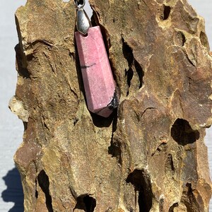 May include: A pink and black gemstone pendant with a silver bail. The pendant is sitting on a rough, brown rock.