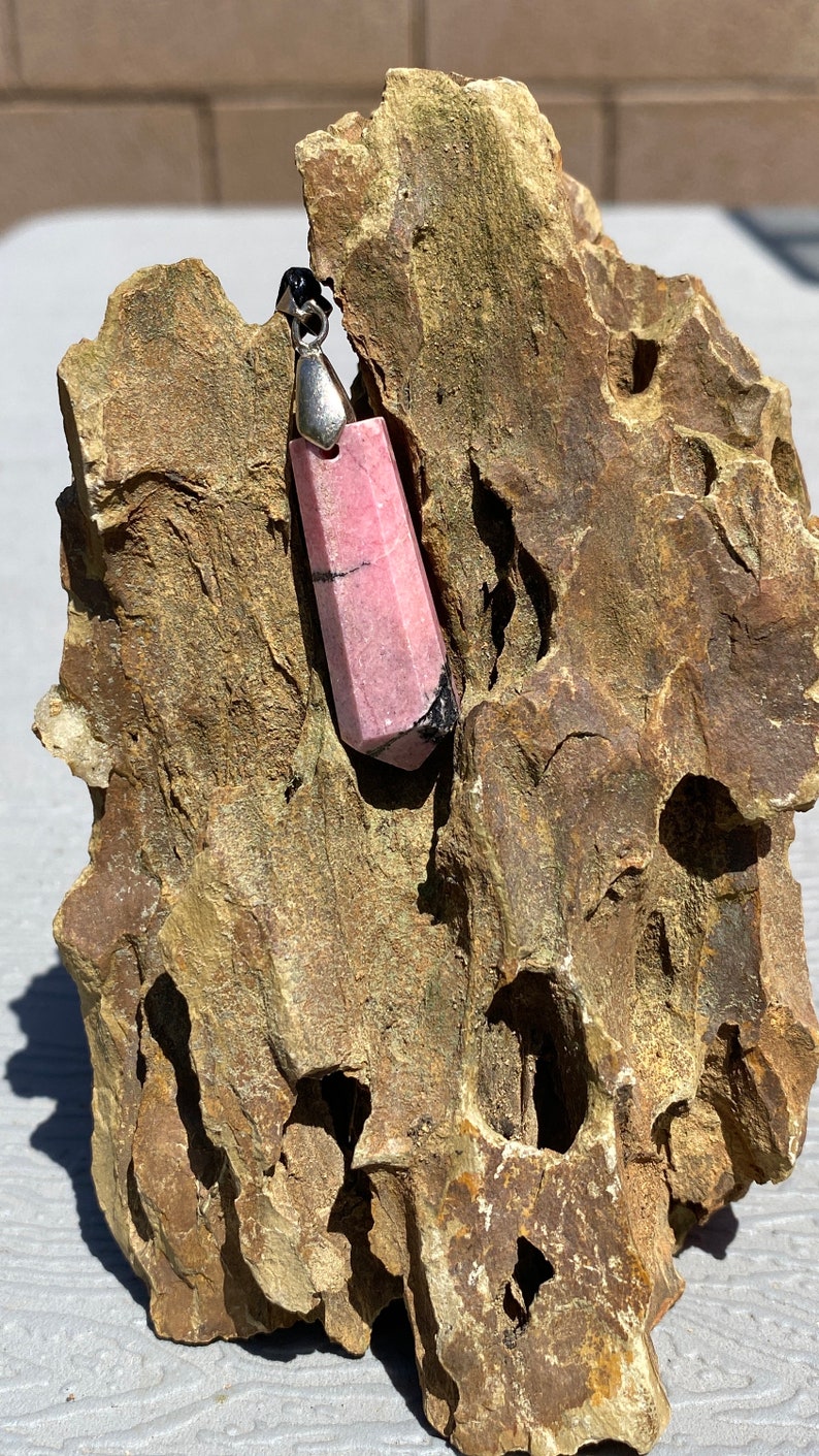 May include: A pink and black gemstone pendant with a silver bail. The pendant is sitting on a rough, brown rock.