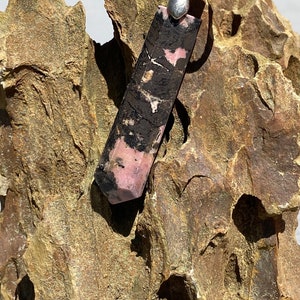 May include: A black and pink gemstone pendant with a silver bail. The pendant is hanging from a black cord and is resting on a piece of rough, brown rock.