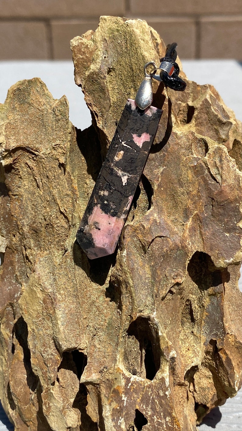 May include: A black and pink gemstone pendant with a silver bail. The pendant is hanging from a black cord and is resting on a piece of rough, brown rock.