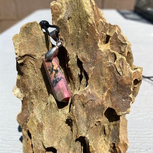 May include: A pink and black gemstone pendant on a black cord. The pendant is a long, rectangular shape with a silver bail.