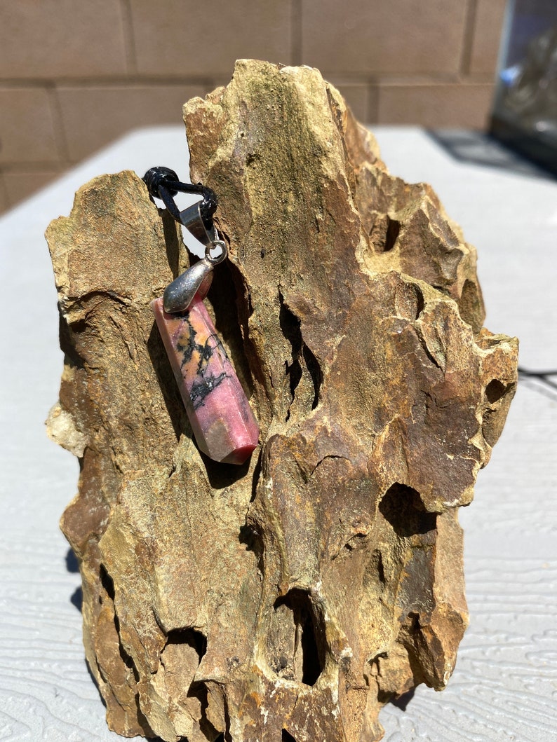 May include: A pink and black gemstone pendant on a black cord. The pendant is a long, rectangular shape with a silver bail.
