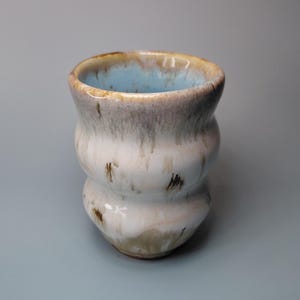 May include: A ceramic cup with a wavy, double-bulge silhouette. The exterior features a glossy white glaze with brown drip-like accents. The interior is finished in a smooth, light blue glaze that extends over the top rim of the vessel.