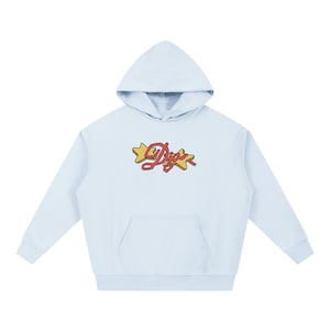 Op de afbeelding: Een lichtblauwe hoodie met een voorzak en een capuchon. De hoodie heeft het woord "Digs" in een cursief lettertype met een rood lint en twee gele steraccenten.