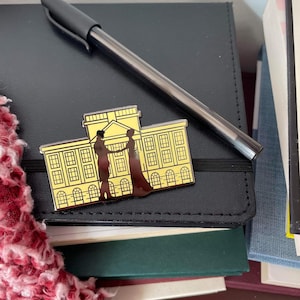 May include: A gold-colored pin featuring a building design with two figures in silhouette, holding hands. The pin is set against a black notebook with a silver pen resting on top. A red and white fuzzy scarf is visible in the corner.