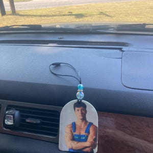 Peut inclure: Un diffuseur d'air pour voiture avec une photo d'un homme portant une chemise bleue et verte et les bras crois&eacute;s. Le diffuseur est suspendu &agrave; une ficelle avec des perles bleues et iris&eacute;es.