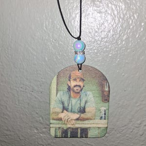 May include: A pendant featuring a printed image of a man wearing a baseball cap and a t-shirt, hanging from a black cord. The cord is adorned with two iridescent blue beads and a silver spacer. The pendant is a light brown color.