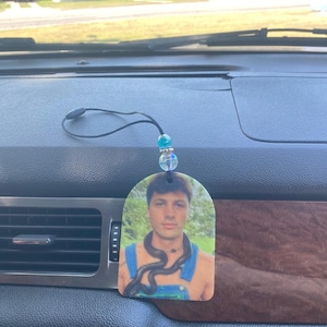 Peut inclure: Un diffuseur d'air pour voiture avec une photo d'une personne avec un serpent autour du cou. Le diffuseur est suspendu &agrave; une ficelle avec des perles bleues et vertes.