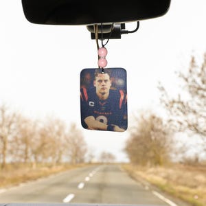 Pode incluir: Um aromatizador de carro com uma foto de um jogador de futebol americano vestindo uma camisa azul e vermelha com o n&uacute;mero 9. O aromatizador est&aacute; pendurado no espelho retrovisor de um carro.