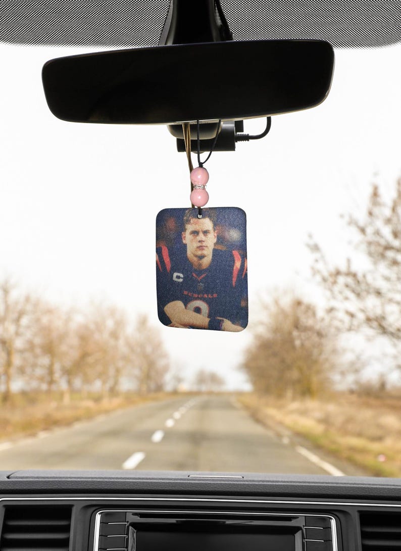 Pode incluir: Um aromatizador de carro com uma foto de um jogador de futebol americano vestindo uma camisa azul e vermelha com o n&uacute;mero 9. O aromatizador est&aacute; pendurado no espelho retrovisor de um carro.