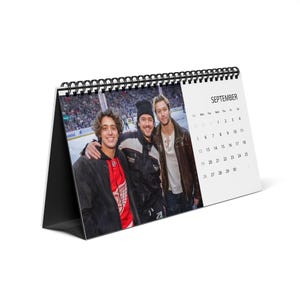 May include: A desk calendar with a black spiral binding featuring a photograph of three people standing together at an ice hockey arena. The right side displays the month of September with a grid of dates printed in black text on a white background.