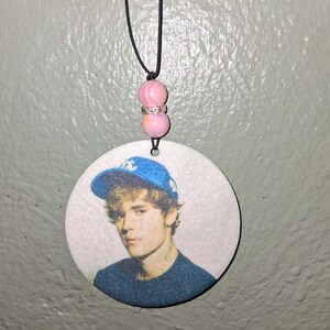 May include: A round pendant featuring a portrait of a person wearing a blue baseball cap. The pendant is suspended by a black cord with pink beads. The person in the portrait is wearing a dark blue shirt.