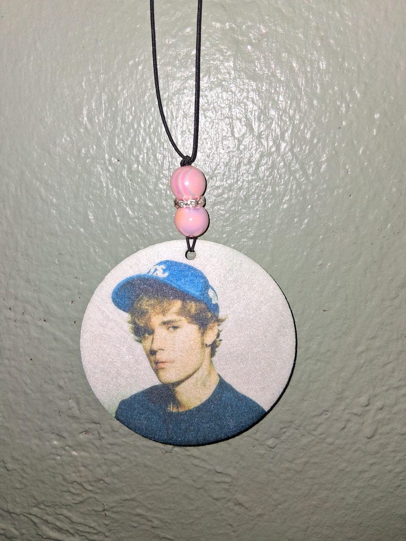 May include: A round pendant featuring a portrait of a person wearing a blue baseball cap. The pendant is suspended by a black cord with pink beads. The person in the portrait is wearing a dark blue shirt.