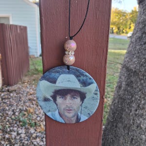 May include: A round pendant necklace featuring a portrait of a man wearing a cowboy hat. The pendant is suspended from a black cord, with two iridescent beads and a silver spacer. The background is a blurred outdoor scene.
