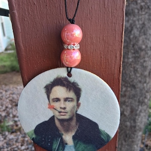 May include: A round, off-white air freshener with a printed image of a man with a red flower in his mouth. The air freshener hangs from a black cord, with two pink beads and a silver spacer.
