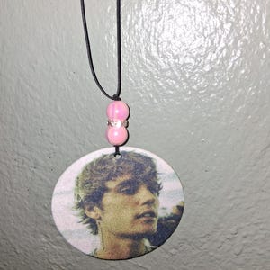 May include: A round pendant necklace featuring a printed image of a person's face. The pendant is suspended from a black cord and has pink beads with a silver spacer. The pendant is a light color.