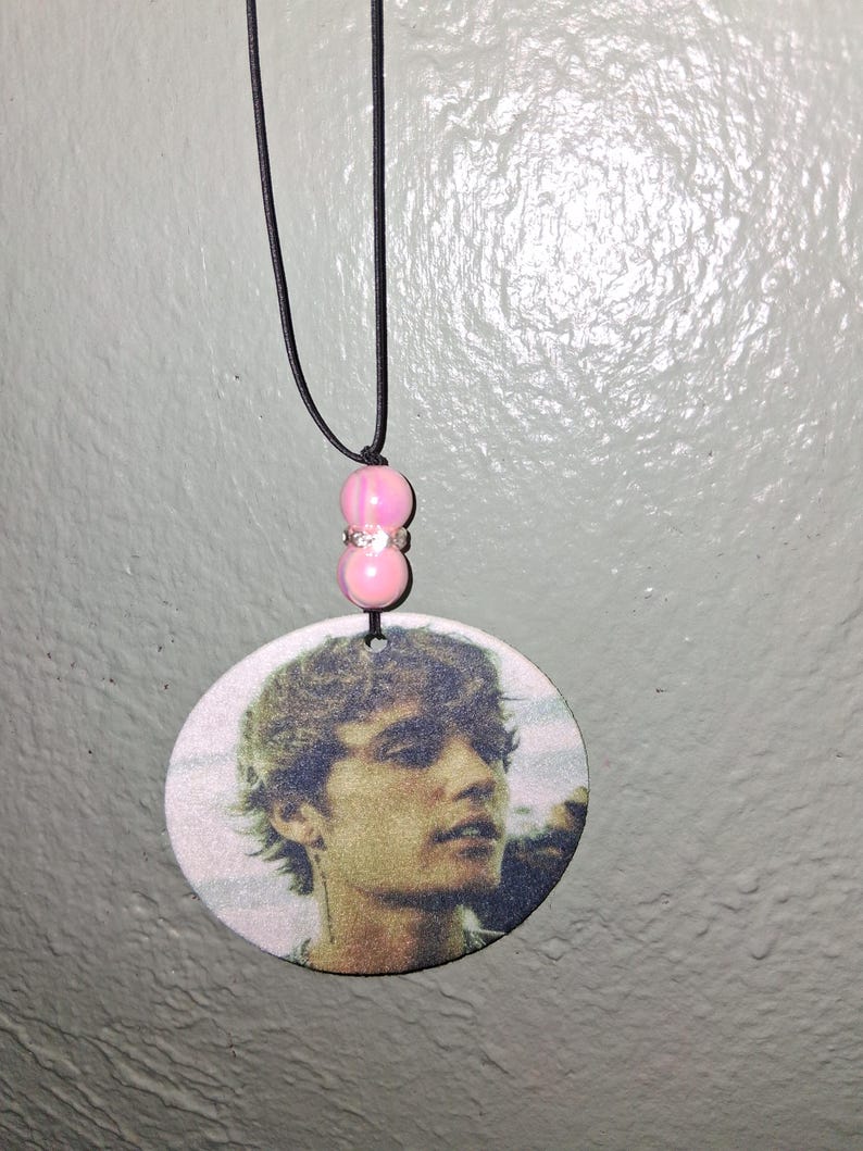 May include: A round pendant necklace featuring a printed image of a person's face. The pendant is suspended from a black cord and has pink beads with a silver spacer. The pendant is a light color.