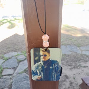 Pode incluir: Um aromatizador de carro com uma imagem de um homem usando &oacute;culos de sol e uma jaqueta. O aromatizador est&aacute; pendurado em um cord&atilde;o preto com duas contas cor-de-rosa.