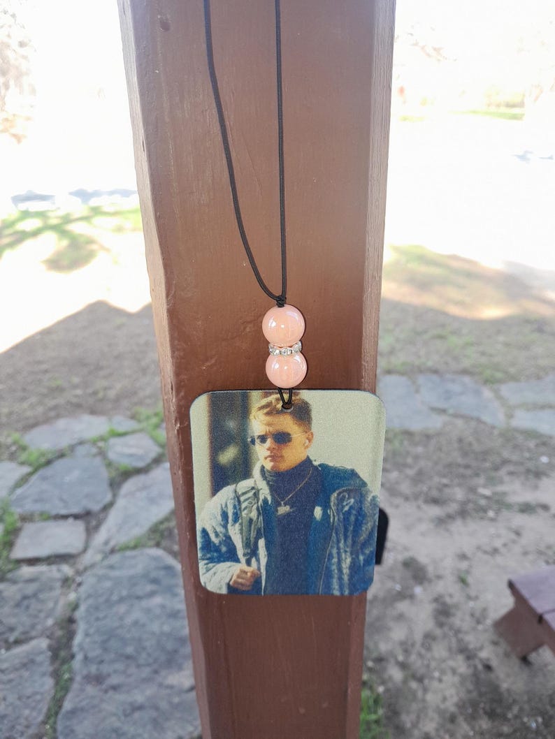 Pode incluir: Um aromatizador de carro com uma imagem de um homem usando &oacute;culos de sol e uma jaqueta. O aromatizador est&aacute; pendurado em um cord&atilde;o preto com duas contas cor-de-rosa.