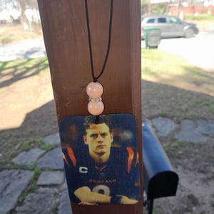 Pode incluir: Um aromatizador de carro com um cord&atilde;o preto e duas contas cor-de-rosa. O aromatizador apresenta uma foto de um jogador de futebol americano vestindo uma camisa azul e laranja com a palavra "Bengals" nela.