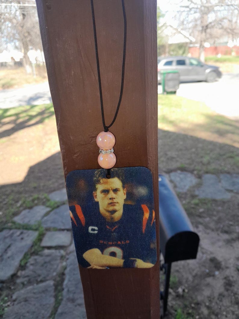 Pode incluir: Um aromatizador de carro com um cord&atilde;o preto e duas contas cor-de-rosa. O aromatizador apresenta uma foto de um jogador de futebol americano vestindo uma camisa azul e laranja com a palavra "Bengals" nela.