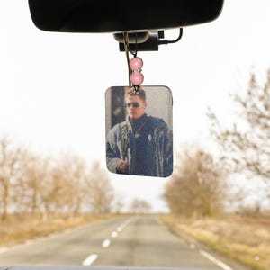 Pode incluir: Um aromatizador de carro com uma foto de um homem usando &oacute;culos de sol e uma jaqueta. O aromatizador est&aacute; pendurado no espelho retrovisor de um carro.