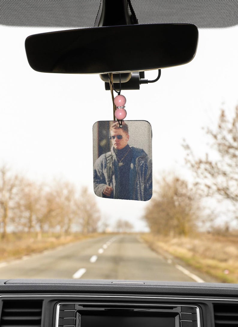 Pode incluir: Um aromatizador de carro com uma foto de um homem usando &oacute;culos de sol e uma jaqueta. O aromatizador est&aacute; pendurado no espelho retrovisor de um carro.