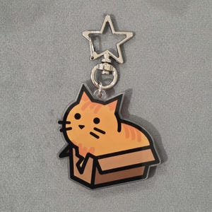 May include: A keyring featuring an orange tabby cat cartoon in a cardboard box. The cat has black whiskers and is smiling. The keyring has a silver star-shaped clasp.