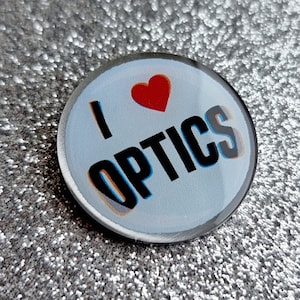 Może przedstawiać: Biały guzik z czerwonym sercem i tekstem "I love OPTICS" czarnymi literami z niebieskim i pomarańczowym obramowaniem.