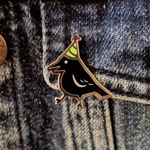 May include: A black crow enamel pin with a silver outline and a party hat. The crow has a white eye and a curved white line on its chest. The party hat is green and yellow. The pin is attached to a denim jacket.