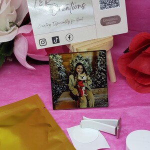 Puede incluir: Un ambientador de coche de cerámica blanca con un círculo adhesivo blanco y un pequeño círculo blanco con un anillo metálico plateado. El ambientador está al lado de una bolsa de papel de aluminio dorado y una foto de una mujer sosteniendo un perro pequeño en una escena de invierno.