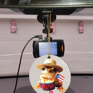 Puede incluir: Un ambientador de coche redondo y blanco con una rana de dibujos animados que lleva un sombrero y sostiene un tambor. La rana sostiene una pequeña bandera puertorriqueña. El ambientador está colgado de un espejo retrovisor en un coche.