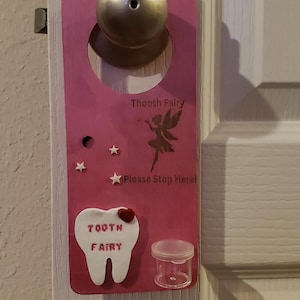 Könnte beinhalten: Rosafarbener Holztürhänger mit einem Zahnfeen-Motiv. Er verfügt über einen silbernen Türknauf, eine ausgeschnittene Fee, einen weißen Zahn mit den Worten "Tooth Fairy" und einen kleinen durchsichtigen Behälter.