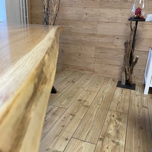 Op de afbeelding: Een close-up van een grote, natuurlijke houten tafel met een levende rand. De tafel is gemaakt van lichtbruin hout met een natuurlijk graanpatroon. De tafel staat op een lichtbruine houten vloer.