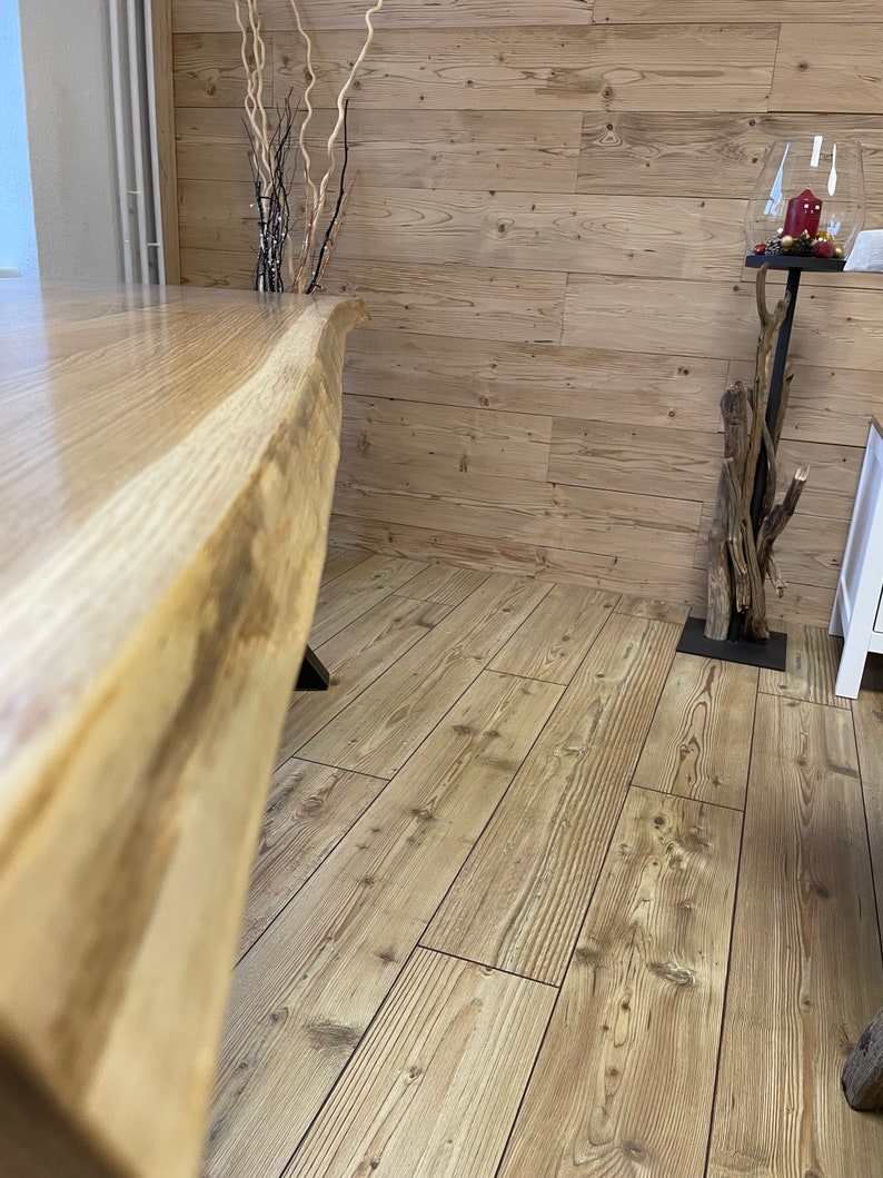 Op de afbeelding: Een close-up van een grote, natuurlijke houten tafel met een levende rand. De tafel is gemaakt van lichtbruin hout met een natuurlijk graanpatroon. De tafel staat op een lichtbruine houten vloer.