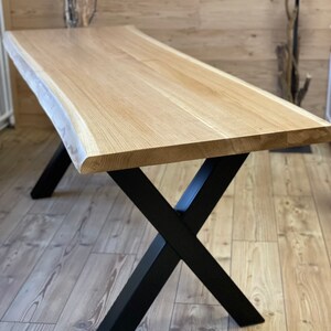 Op de afbeelding: Een houten tafel met een live edge blad en zwarte metalen X-vormige poten. De tafel is gemaakt van natuurlijk hout en heeft een rustieke uitstraling.