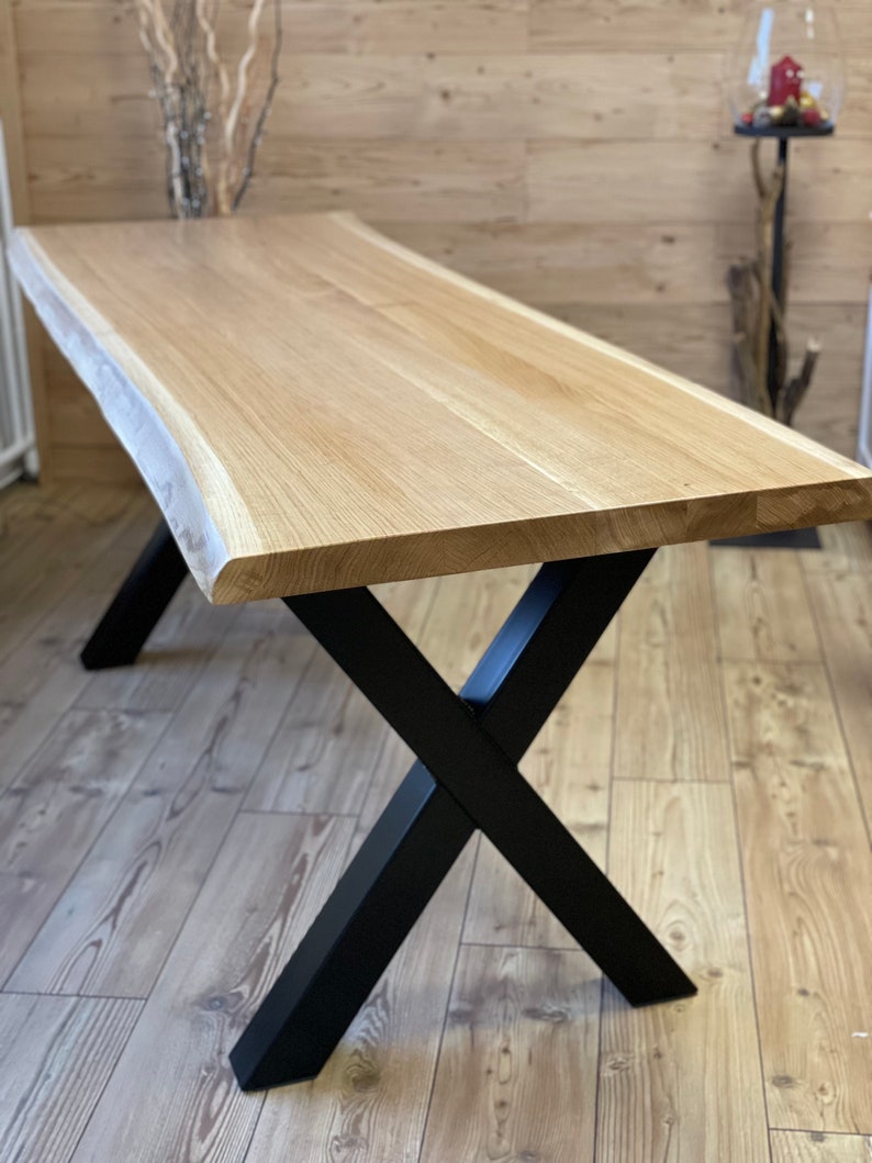 Op de afbeelding: Een houten tafel met een live edge blad en zwarte metalen X-vormige poten. De tafel is gemaakt van natuurlijk hout en heeft een rustieke uitstraling.