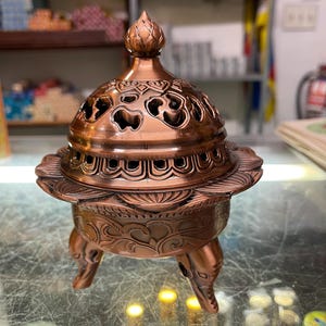 May include: A copper-colored incense burner with intricate floral and geometric cut-out designs. The burner has a domed lid with a decorative finial and sits on three ornate legs. The base features detailed relief patterns.