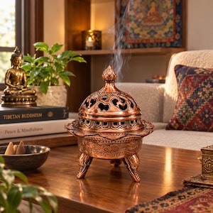 May include: A copper-colored metal incense burner with a dome-shaped lid featuring intricate cutouts and a finial top. The burner stands on three legs and emits a thin trail of smoke from the top, showcasing detailed embossed patterns.