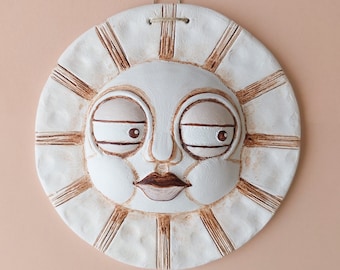 May include: White ceramic sun wall hanging with a brown painted design. The sun has a face with closed eyes, a nose, and a mouth.