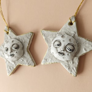 Peut inclure: Deux ornements en forme d'étoile sur le thème céleste. Chaque ornement présente un visage ressemblant à la lune avec les yeux fermés. Les étoiles sont blanches avec une bordure dorée et une ficelle dorée pour suspendre.