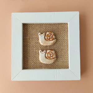 May include: Two ceramic snail ornaments with brown and cream-coloured shells and smiling faces are displayed on a burlap background within a white wooden frame. The frame has a square shape.