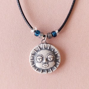 May include: A white ceramic sun pendant necklace with a face and closed eyes. The necklace has a black cord and blue and silver beads.