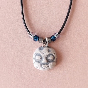 May include: A handmade necklace featuring a pendant with a face design. The pendant is a light gray color with darker gray details. The necklace has a black cord with small beads in shades of blue, silver, and pink.