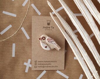 May include: A ceramic bird brooch in cream and brown, with decorative details. The brooch is on a brown card with the text "Indre Ta STUDIO". The background includes dried plant stems and a brown paper with white markings.