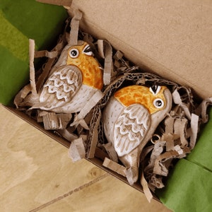May include: Two ceramic bird ornaments in a box. The birds are white with brown and orange accents. They are sitting on a bed of brown packing peanuts.