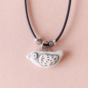 May include: A ceramic bird pendant necklace with a black cord and silver beads. The bird is white with gray markings and has a small eye.